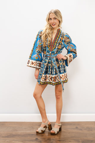 Woman wearing a colorful patterned dress standing against a white wall.