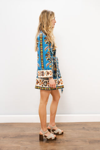Woman wearing a colorful patterned dress standing on a wooden floor with a white wall background
