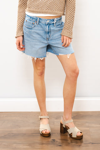 Person wearing light blue denim shorts and beige sandals on a wooden floor.