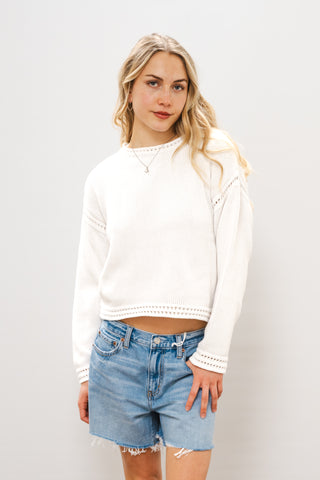 woman in a white sweater and blue denim shorts standing against a blank wall