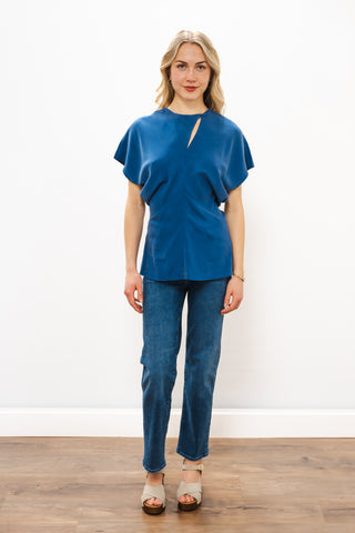 Woman wearing a blue top and jeans standing on a wooden floor with a white wall background