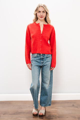 Woman wearing a red cardigan and blue jeans against a white wall.