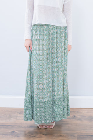 Person wearing a long green patterned skirt with a white top on a wooden floor.