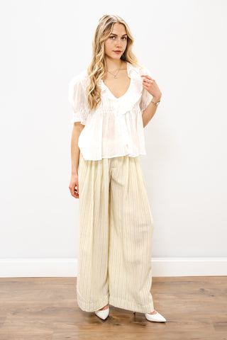 Woman wearing a white blouse and beige striped pants against a white wall.