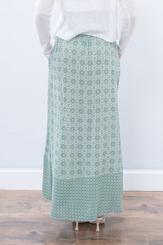 Person wearing a long green patterned skirt on a wooden floor with a white wall background