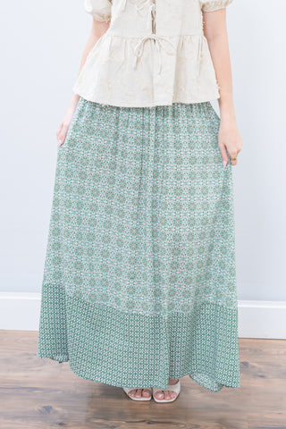 Person wearing a long green patterned skirt and beige top against a white wall.