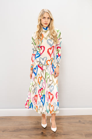 a woman standing in white heels against a white wall in a multi colorful long sleeve turtleneck and skirt