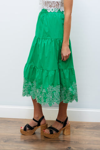 Woman wearing a white lace top and green tiered skirt against a white wall.