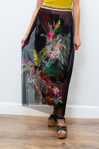 Woman wearing a yellow top and floral sheer skirt against a white wall.