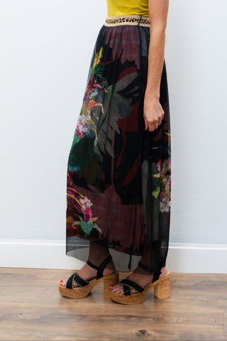 Woman wearing a yellow top and a long, dark floral skirt against a white wall.