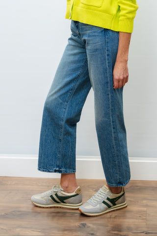 Woman wearing a bright yellow jacket and blue jeans standing against a white wall.